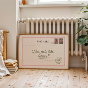 May include: A framed postcard print with a light pink border and the words "This feels like home" in a handwritten style. The print includes postage stamps and a postmark design. The frame is a light brown color, and the print is displayed on a wooden floor.