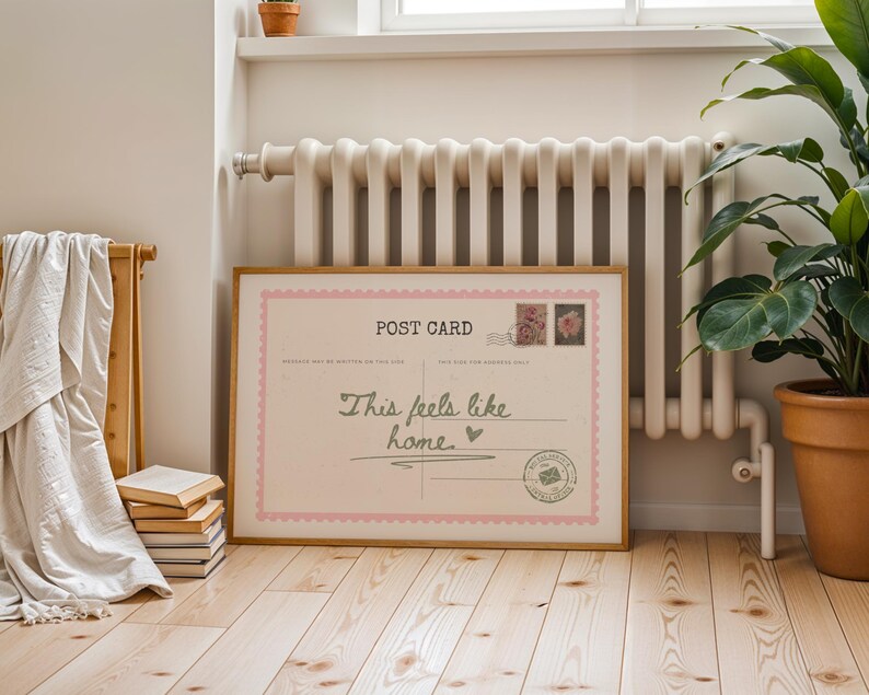 May include: A framed postcard print with a light pink border and the words "This feels like home" in a handwritten style. The print includes postage stamps and a postmark design. The frame is a light brown color, and the print is displayed on a wooden floor.