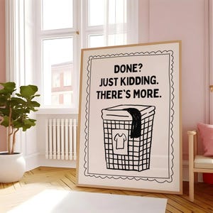 May include: A framed print with the text "DONE? JUST KIDDING. THERE'S MORE." above a black and white illustration of a laundry basket with a shirt and a dark cloth. The print has a light brown frame and is displayed in a room with a window.
