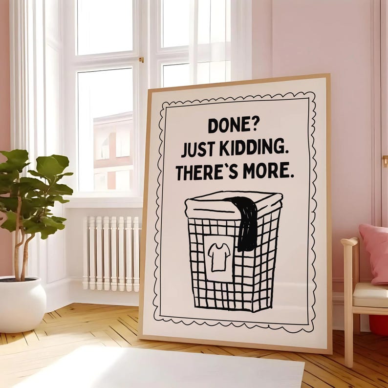 May include: A framed print with the text "DONE? JUST KIDDING. THERE'S MORE." above a black and white illustration of a laundry basket with a shirt and a dark cloth. The print has a light brown frame and is displayed in a room with a window.