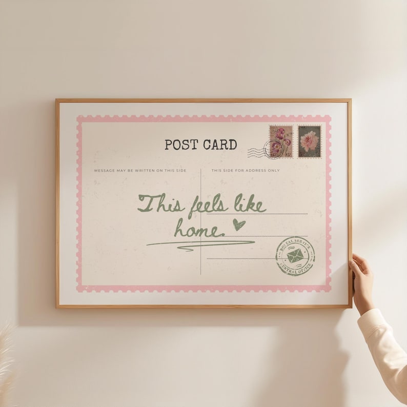 May include: A framed postcard print with a light pink border and the words "POST CARD" at the top. The text "This feels like home" is written in cursive. Two postage stamps and a postal service seal are also visible.