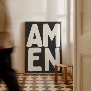May include: A framed artwork with the word "AMEN" in large, white, bold letters against a black background. The artwork is in a room with a checkered floor and a small wooden stool.