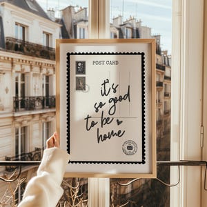 May include: A framed postcard print with the text "it's so good to be home" in a handwritten style. The postcard has a black border and postage stamp illustrations. The frame is a light wood color.