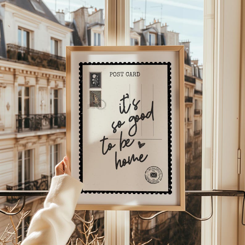 May include: A framed postcard print with the text "it's so good to be home" in a handwritten style. The postcard has a black border and postage stamp illustrations. The frame is a light wood color.