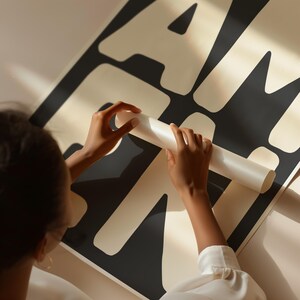 May include: A large black and cream-colored poster with bold, abstract lettering. A person is holding a rolled-up section of the poster. The poster is partially unrolled, revealing the design. The overall aesthetic is modern and minimalist.