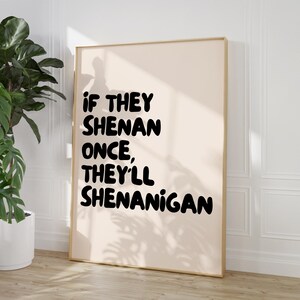 May include: A framed print with the text "IF THEY SHENAN ONCE, THEY'LL SHENANIGAN" in bold black letters. The background is a light beige color, and the frame is a light brown color. The print is leaning against a white wall.