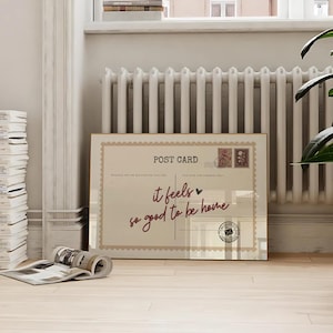 May include: A large rectangular postcard with the words "it feels so good to be home" in a handwritten font. The postcard has a vintage design with postage stamps and a postmark. The postcard is leaning against a white radiator.