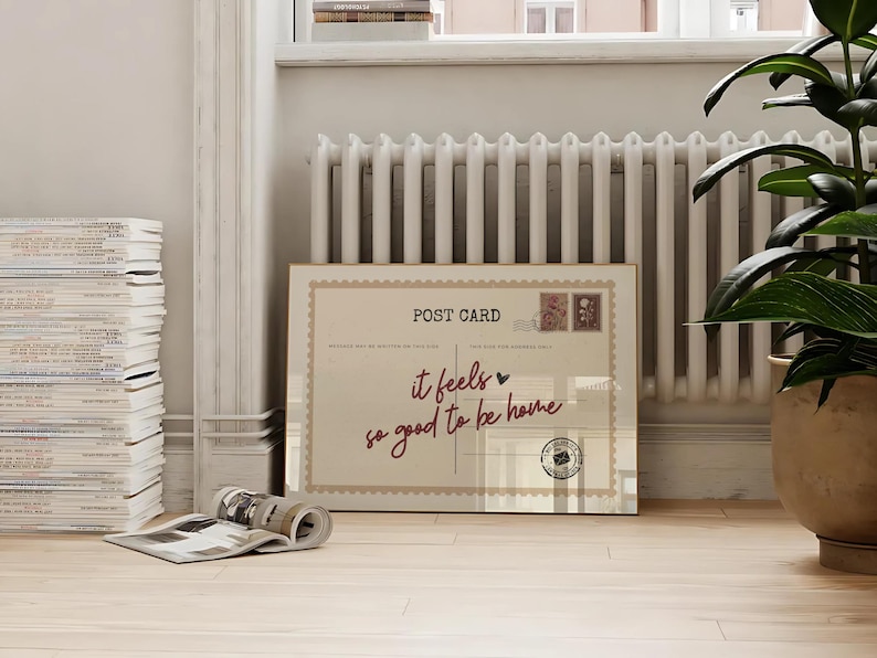 May include: A large rectangular postcard with the words "it feels so good to be home" in a handwritten font. The postcard has a vintage design with postage stamps and a postmark. The postcard is leaning against a white radiator.