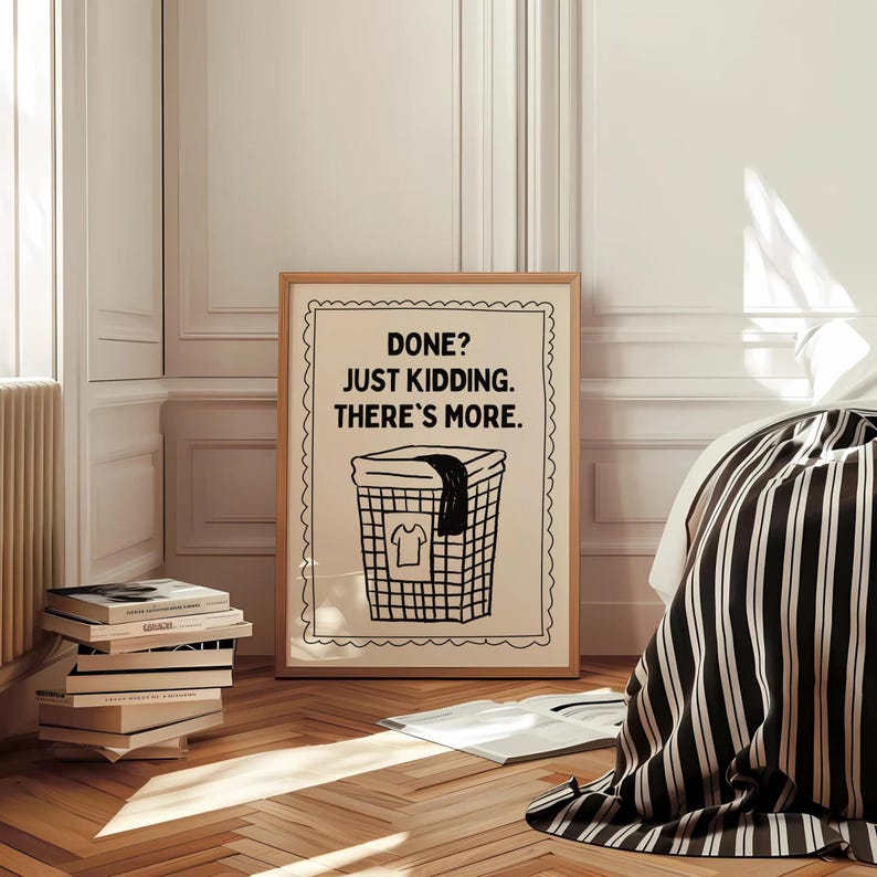 May include: A framed print with the text "DONE? JUST KIDDING. THERE'S MORE." above a laundry basket illustration. The artwork is in a room with a bed, books, and wooden flooring.