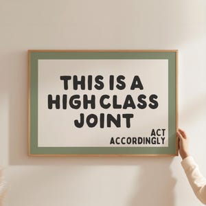 May include: A framed art print with the text "THIS IS A HIGH CLASS JOINT ACT ACCORDINGLY" in bold black letters. The print has a light green border and a natural wood frame, displayed against a white wall.