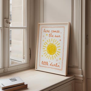 May include: A framed print with the words "here comes the sun little darlin" in orange script. The print features a yellow sun with rays and stars on a white background, set in a light wood frame. The print is displayed on a windowsill.