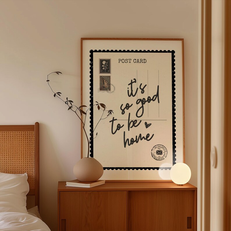 May include: A framed postcard print with the text "it's so good to be home" in black script. The print is displayed above a wooden cabinet, with a beige vase and a small, round lamp. A sprig of greenery adds a natural touch.