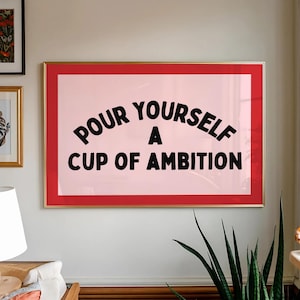 May include: A framed print with the text "POUR YOURSELF A CUP OF AMBITION" in black, arched lettering on a pink background. The frame is red with a gold border. The print is hanging on a white wall.