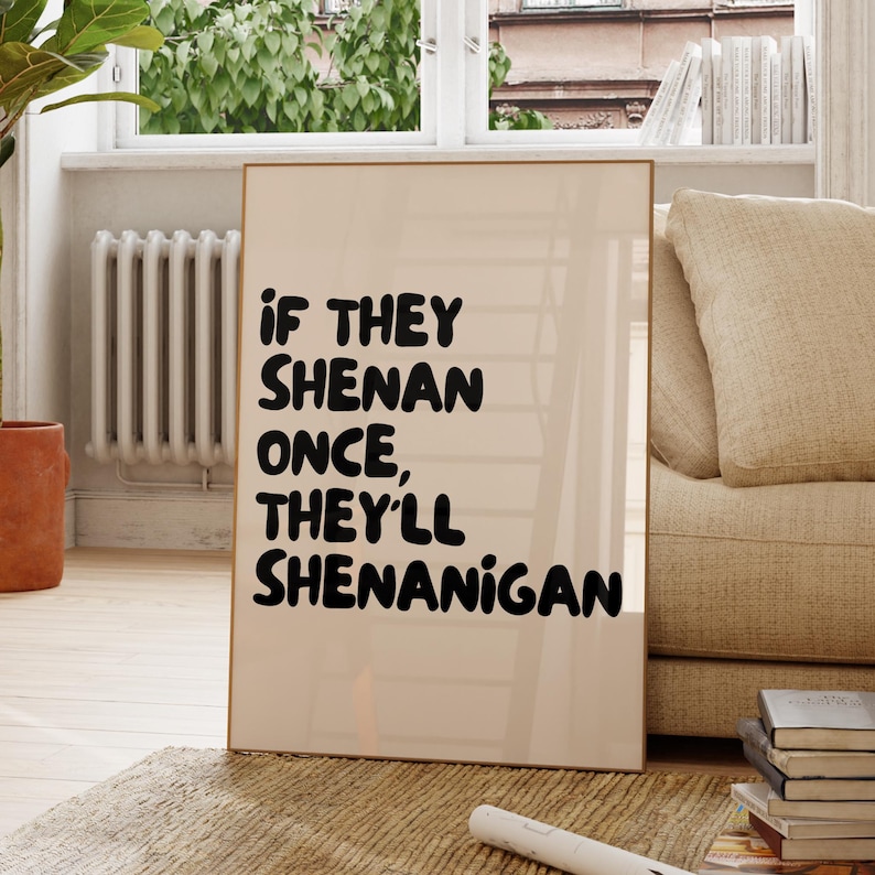 May include: A framed print with the text "If they shenan once, they'll shenanigan" in a bold, black font. The print has a light beige background and a thin, wooden frame. The print is displayed in a living room setting.
