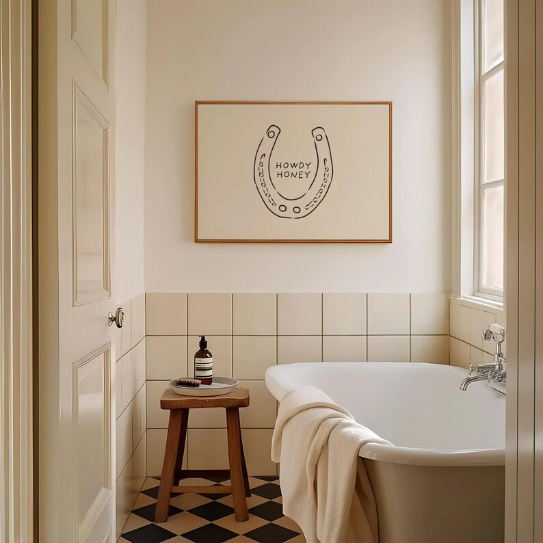 May include: A bathroom scene featuring a framed artwork with a horseshoe design and the words "HOWDY HONEY". The room includes a white bathtub, a wooden stool, and a black and white checkered floor. A bottle of soap and a brush are on the stool.