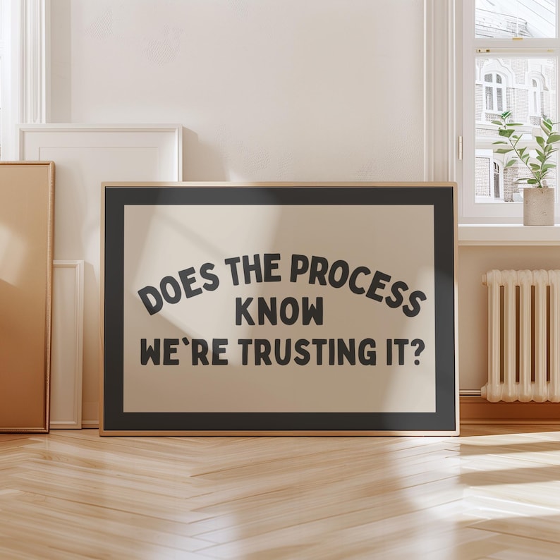 May include: A framed art print with the text "DOES THE PROCESS KNOW WE'RE TRUSTING IT?" in black, arched over a cream-coloured background. The frame is black. The print is displayed indoors, leaning against a wall.