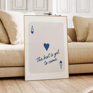 May include: Framed art print featuring a playing card design. The card is white with blue heart symbols and the text "The best is yet to come" in a cursive font. The frame is gold.