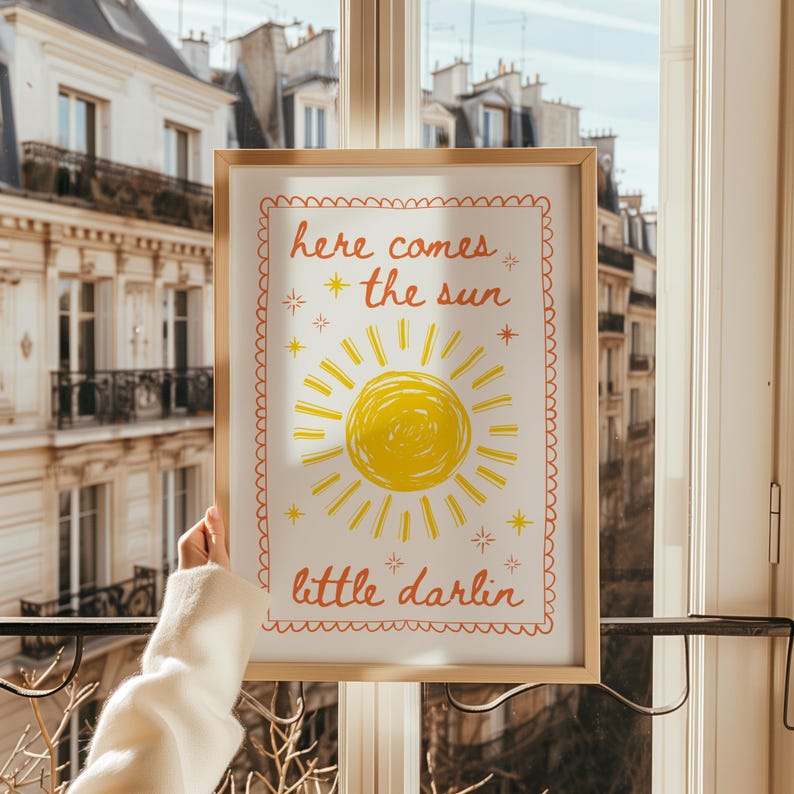 May include: A framed art print with a yellow sun illustration and the words "here comes the sun" and "little darlin" in orange script. The artwork is in a light wood frame, held up in front of a window.