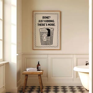 May include: A framed print in a bathroom setting. The artwork features a black and white illustration of a laundry basket with the text "DONE? JUST KIDDING. THERE'S MORE." A wooden stool with a towel and soap bottle is in front of the wall.