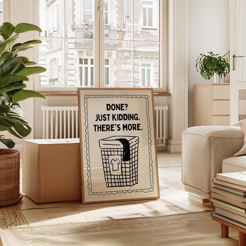 May include: A framed print with the text "DONE? JUST KIDDING. THERE'S MORE." above a black and white illustration of a laundry basket. The print is in a light-colored wooden frame, set against a neutral-toned interior with a large window.