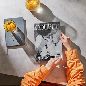 May include: A person holding a magazine titled "COUPLE MAGAZINE" featuring Selena & Justin on the cover. The image is in black and white. A glass of orange-infused drink and a blue notebook are also visible.