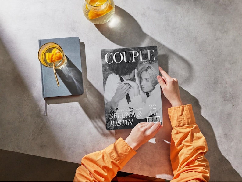 May include: A person holding a magazine titled "COUPLE MAGAZINE" featuring Selena & Justin on the cover. The image is in black and white. A glass of orange-infused drink and a blue notebook are also visible.