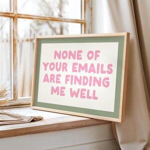 May include: A framed print with the text "NONE OF YOUR EMAILS ARE FINDING ME WELL" in pink letters. The print has a green border and a natural wood frame, resting on a wooden surface near a window.