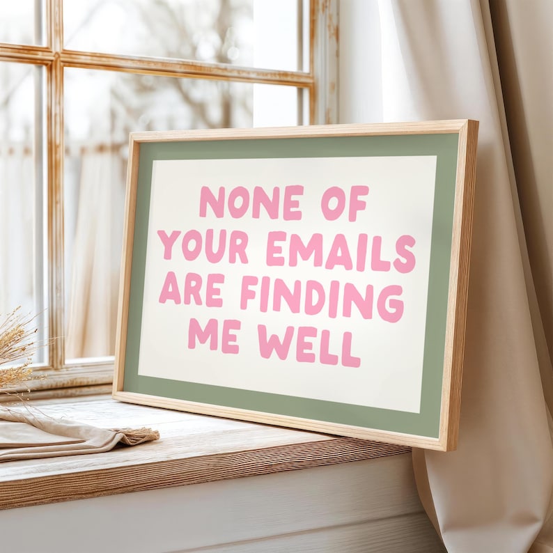 May include: A framed print with the text "NONE OF YOUR EMAILS ARE FINDING ME WELL" in pink letters. The print has a green border and a natural wood frame, resting on a wooden surface near a window.