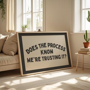 May include: A framed art print with the text "DOES THE PROCESS KNOW WE'RE TRUSTING IT?" in black on a beige background. The frame is black with a natural wood border. The print is leaning against a beige sofa in a well-lit room.