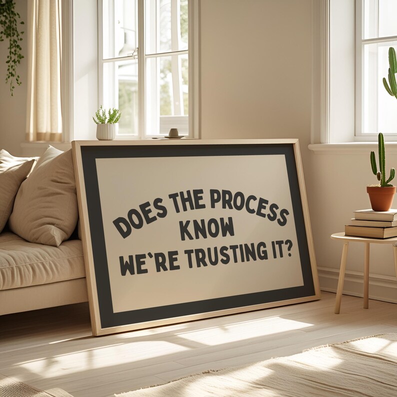 May include: A framed art print with the text "DOES THE PROCESS KNOW WE'RE TRUSTING IT?" in black on a beige background. The frame is black with a natural wood border. The print is leaning against a beige sofa in a well-lit room.