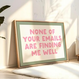 May include: A framed art print with the text "NONE OF YOUR EMAILS ARE FINDING ME WELL" in pink letters. The print has a white background, a green border, and a natural wood frame. The artwork is leaning against a wall.
