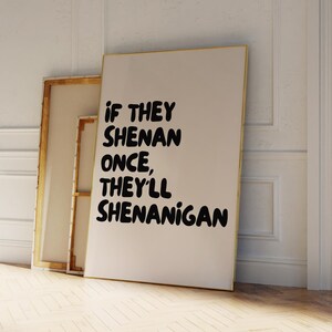 May include: A framed art print with the text "IF THEY SHENAN ONCE, THEY'LL SHENANIGAN" in black, bold letters. The print is displayed against a white wall and wooden floor. Two other framed canvases are leaning against the wall.