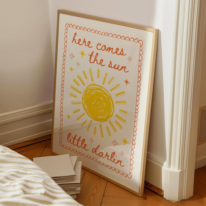 May include: A framed art print with a cheerful design. The artwork features a yellow sun with rays and the words "here comes the sun little darlin" in orange script. The print has a white background and a decorative orange border.