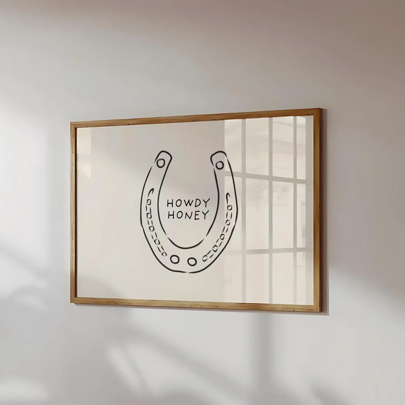 May include: A framed print featuring a black horseshoe design with the words "HOWDY HONEY" in the center. The artwork is set against a cream-colored background and encased in a wooden frame, suitable for wall decor.