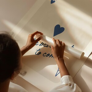 May include: A large, off-white poster with blue heart shapes and the handwritten text "the best is to come." The poster is being unrolled by a person, revealing the message. The overall aesthetic is clean and modern.