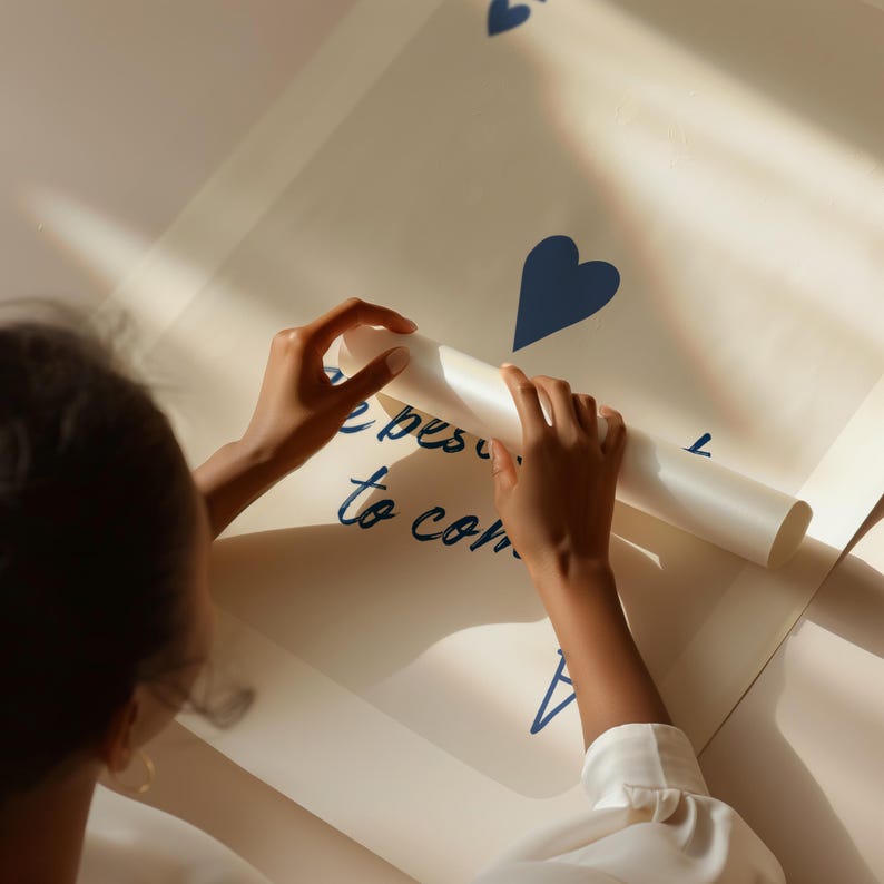 May include: A large, off-white poster with blue heart shapes and the handwritten text "the best is to come." The poster is being unrolled by a person, revealing the message. The overall aesthetic is clean and modern.
