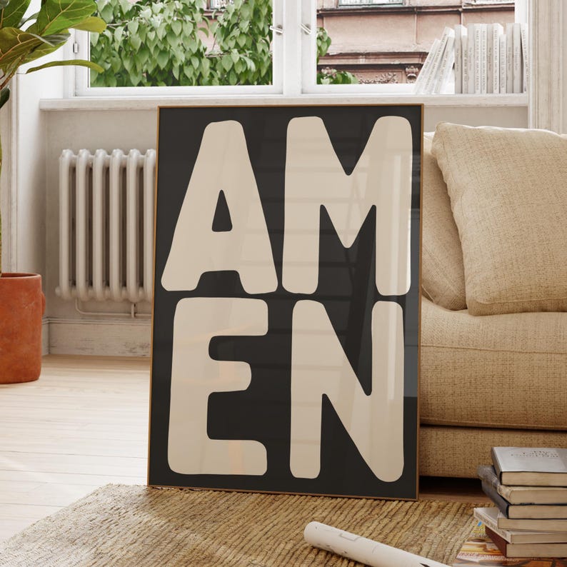 May include: A framed art print with the word "AMEN" in large, bold, off-white letters against a dark gray background. The artwork is displayed in a living room setting, leaning against a wall.