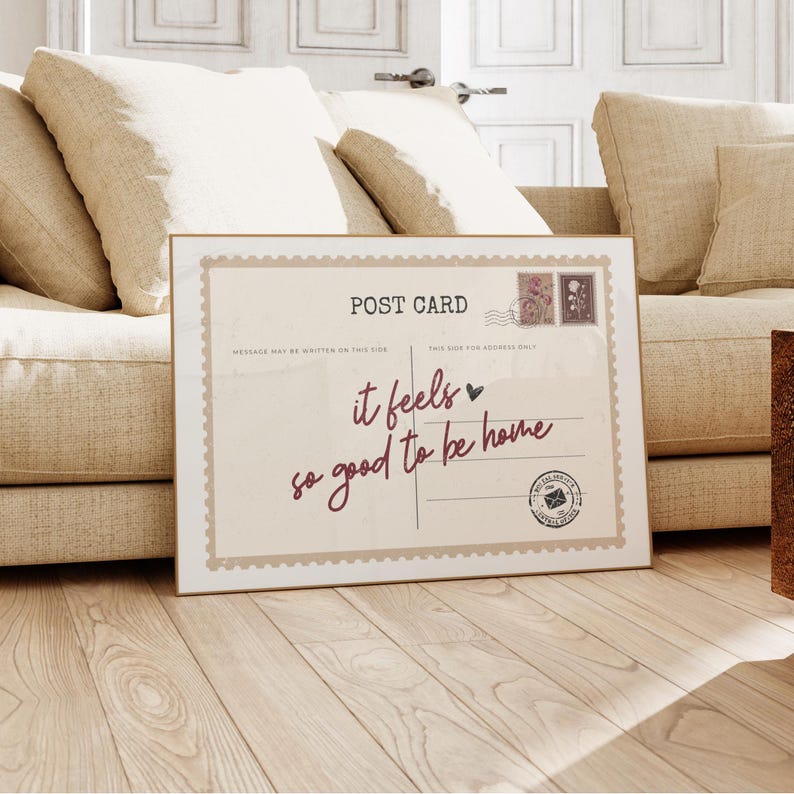 May include: A framed postcard print with the words "it feels so good to be home" in burgundy script. The print has a vintage aesthetic with postage stamp illustrations and a postmark seal. The frame is a light brown color.
