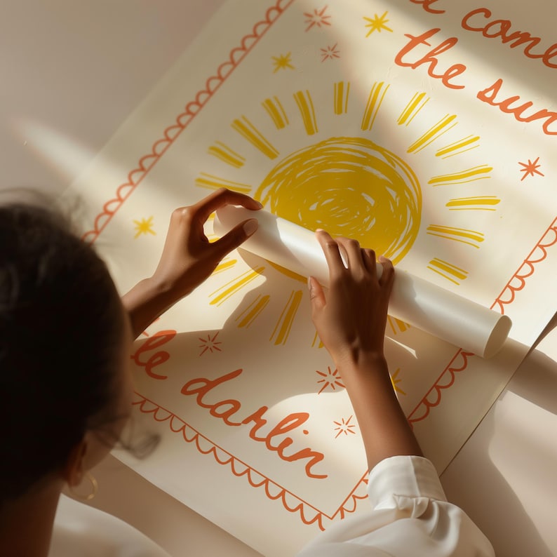 May include: A rolled-up poster with a yellow sun illustration and orange text that reads "come the sun" and "little darlin" on a cream background. The poster is being unrolled by a person.