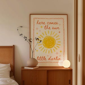 May include: A framed poster with the text "here comes the sun" and "little darlin" in orange and yellow. The poster features a yellow sun illustration. A small, round, white lamp sits on a wooden cabinet.