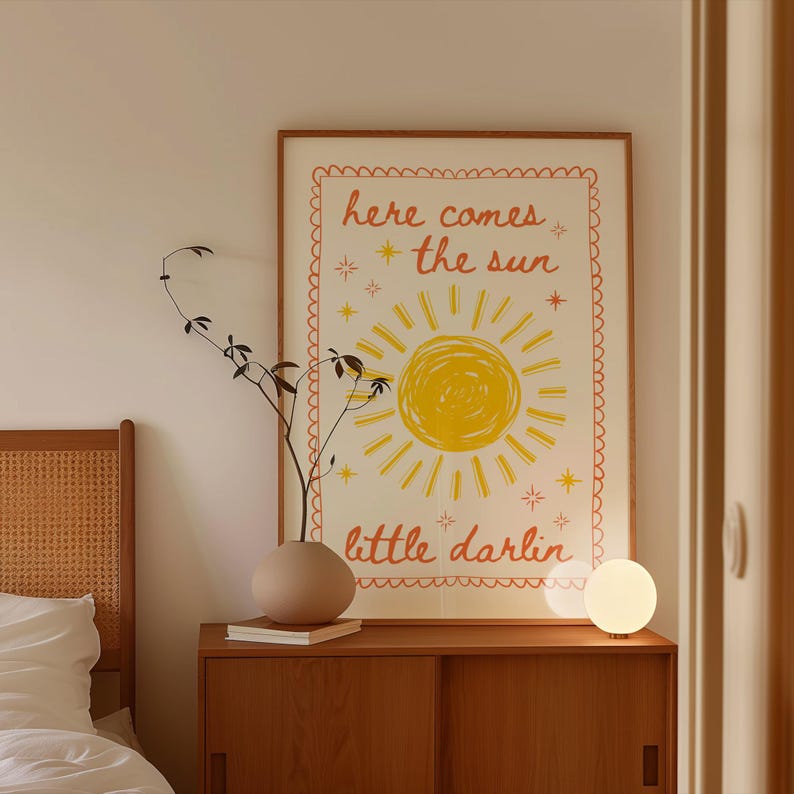 May include: A framed poster with the text "here comes the sun" and "little darlin" in orange and yellow. The poster features a yellow sun illustration. A small, round, white lamp sits on a wooden cabinet.