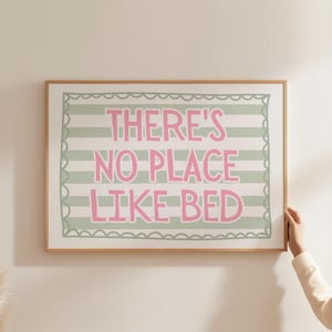 May include: A framed print with the text "THERE'S NO PLACE LIKE BED" in pink letters. The background features horizontal stripes in light green and white. The frame is made of light brown wood.