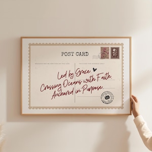 May include: A framed postcard print with a light beige background and a wooden frame. The postcard features the words "Led by Grace, Crossing Oceans with Faith, Anchored in Purpose" in a handwritten style. Two postage stamps are visible.