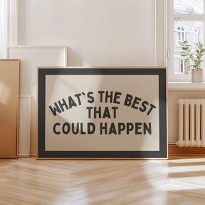 May include: A framed print with the text "WHAT'S THE BEST THAT COULD HAPPEN" in black, arched over a cream-colored background. The frame is black and the print is displayed in a room with a window and wooden floor.