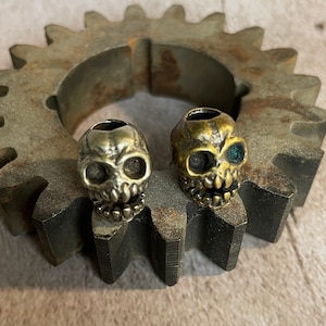 May include: Two small gold skull beads with black eyes. The skull on the right has a green eye. The beads are sitting on a rusty metal gear.