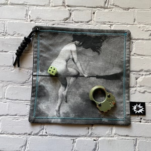 Puede incluir: Una bolsa gris y negra con ribete azul. La bolsa tiene un cordón de ajuste negro y un gráfico de una mujer montando una escoba. Hay un dado verde y un anillo de metal verde en la bolsa.