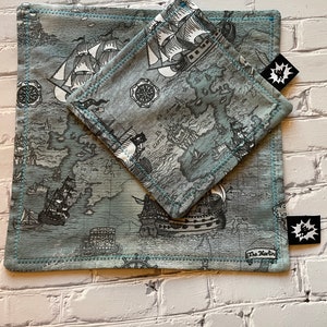 Może przedstawiać: Dwuczęściowy zestaw z motywem mapy z żaglowcami w odcieniach szarości i bieli na tle w kolorze morskim. Zestaw zawiera kwadratowy przedmiot i mniejszy, złożony kwadratowy przedmiot, oba z niebieskimi szwami. Dołączona jest czarna metka z białą grafiką.