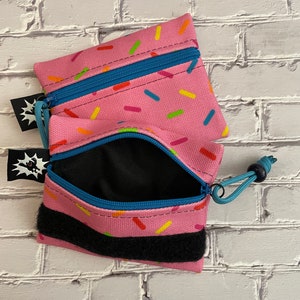 May include: Two pink zippered pouches with a sprinkle pattern. The pouches have a black velcro strap and a blue zipper pull. The pouches are open and the inside is black.