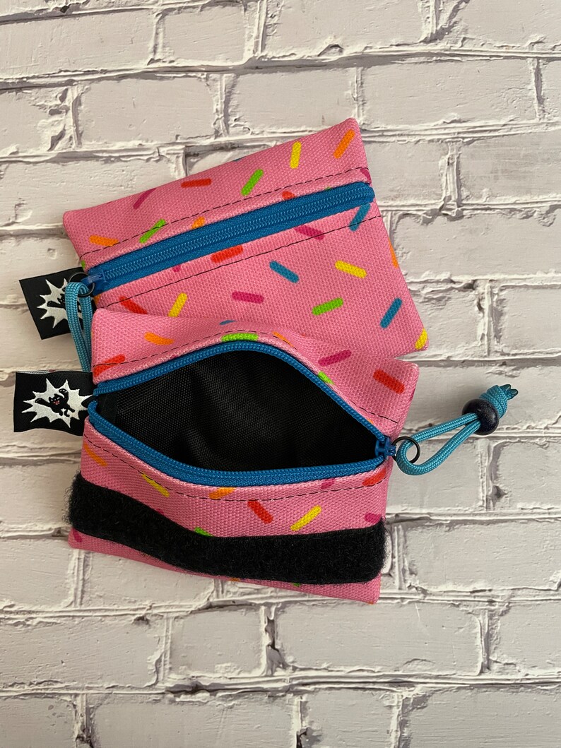 May include: Two pink zippered pouches with a sprinkle pattern. The pouches have a black velcro strap and a blue zipper pull. The pouches are open and the inside is black.