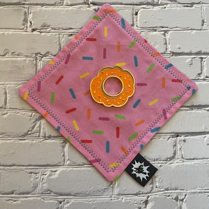 Könnte beinhalten: Ein rosa Stoffquadrat mit einer Donut-förmigen Anstecknadel in der Mitte. Der Stoff hat einen weißen Hintergrund mit bunten Streuseln. Die Anstecknadel ist golden mit orangefarbenem Zuckerguss und weißen Streuseln. Der Stoff hat einen gestickten Rand.